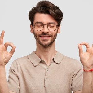 Portrait of happy unshaven young male hipster makes ok sign, has stubble, keeps eyes closed, meditates indoor, poses against white background. Glad Caucasian man gestures indoor and shows approval